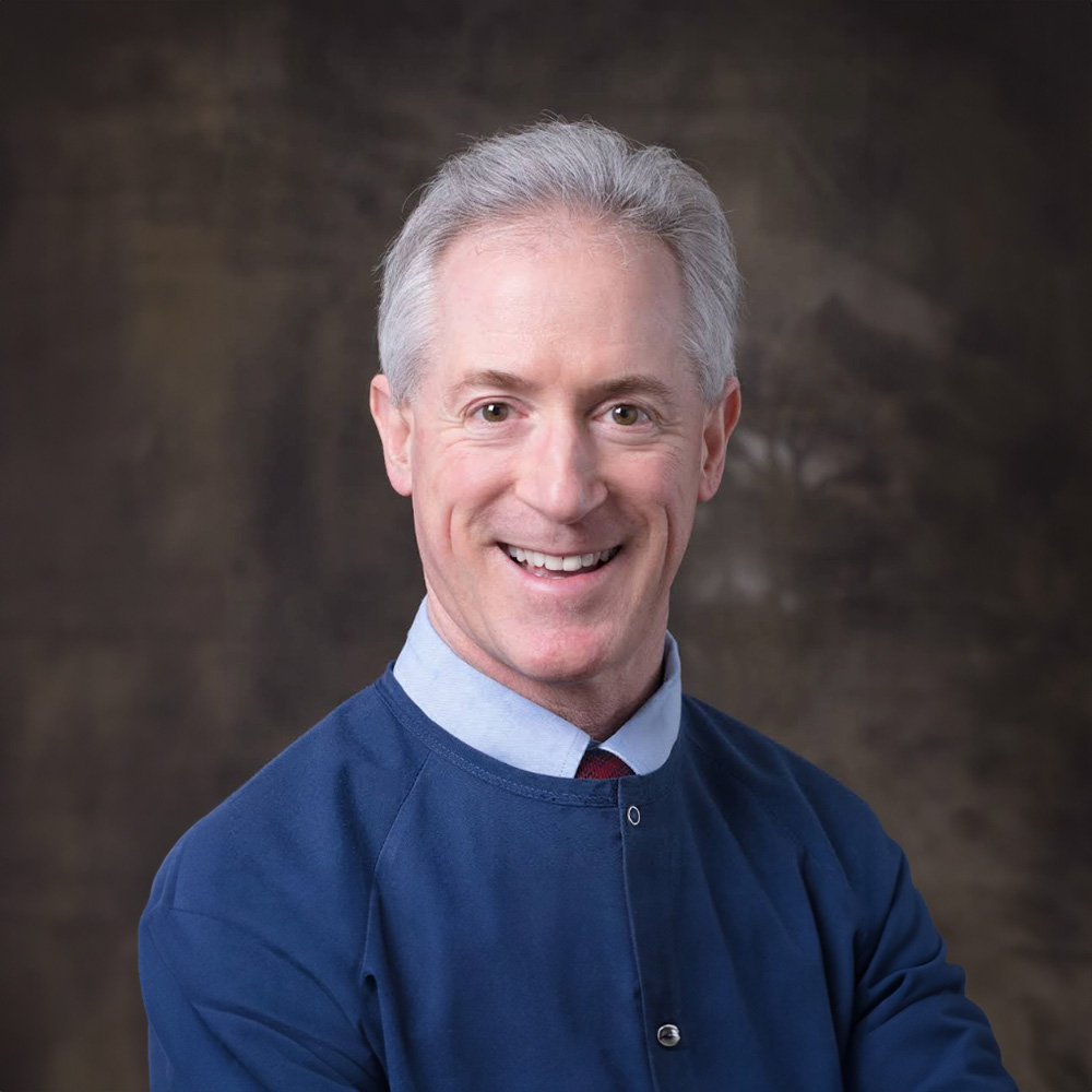 The image shows a man with a smile, wearing a blue sweater, standing against a dark background with a textured surface.