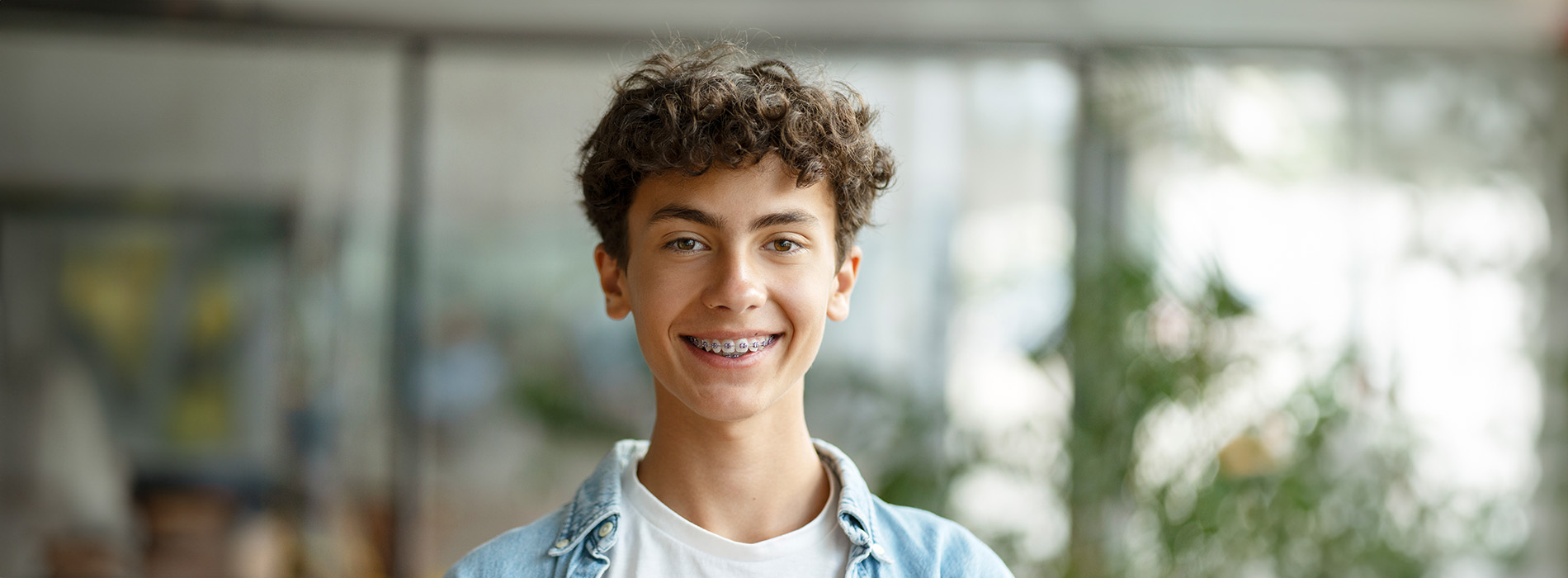 The image shows a young individual with short hair smiling at the camera, wearing a blue jacket and standing indoors against a blurred background.