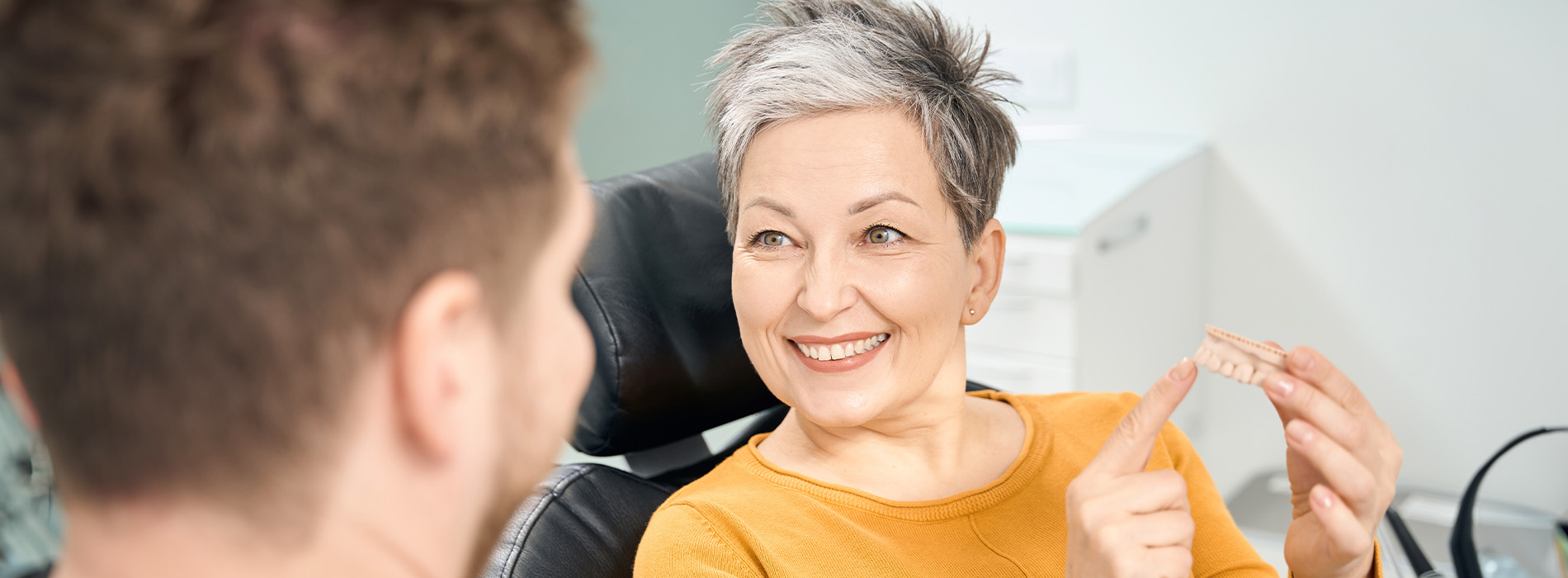 The image shows a person with gray hair sitting in front of another individual who appears to be taking a photo. They are both indoors, possibly in an office setting, with a blurred background that suggests a professional environment.