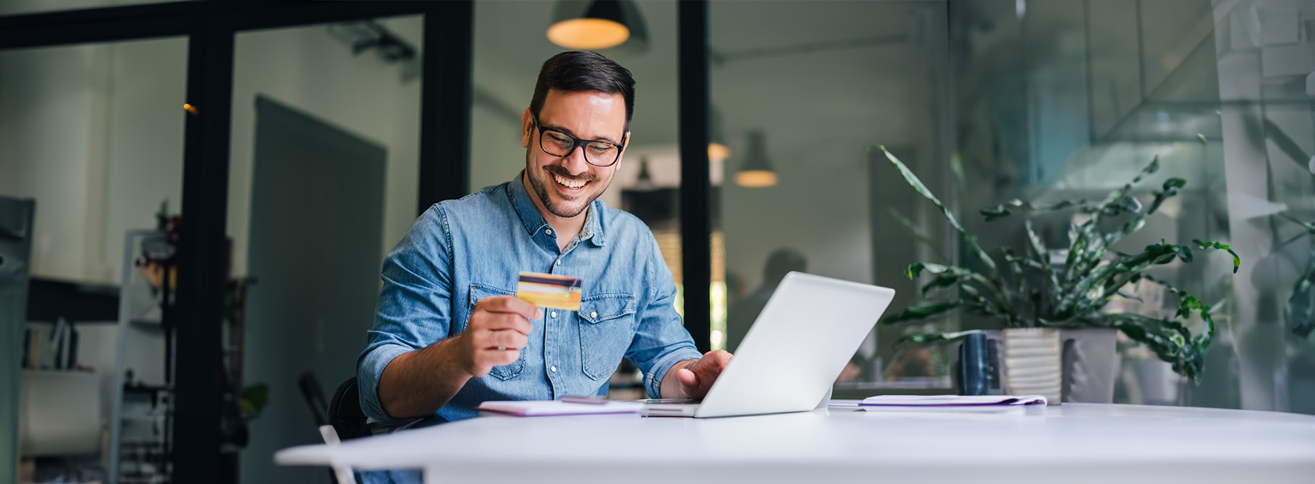 The image shows a man seated at a desk holding a card with his face on it, smiling and looking towards the camera, against a blurred background of an indoor setting.