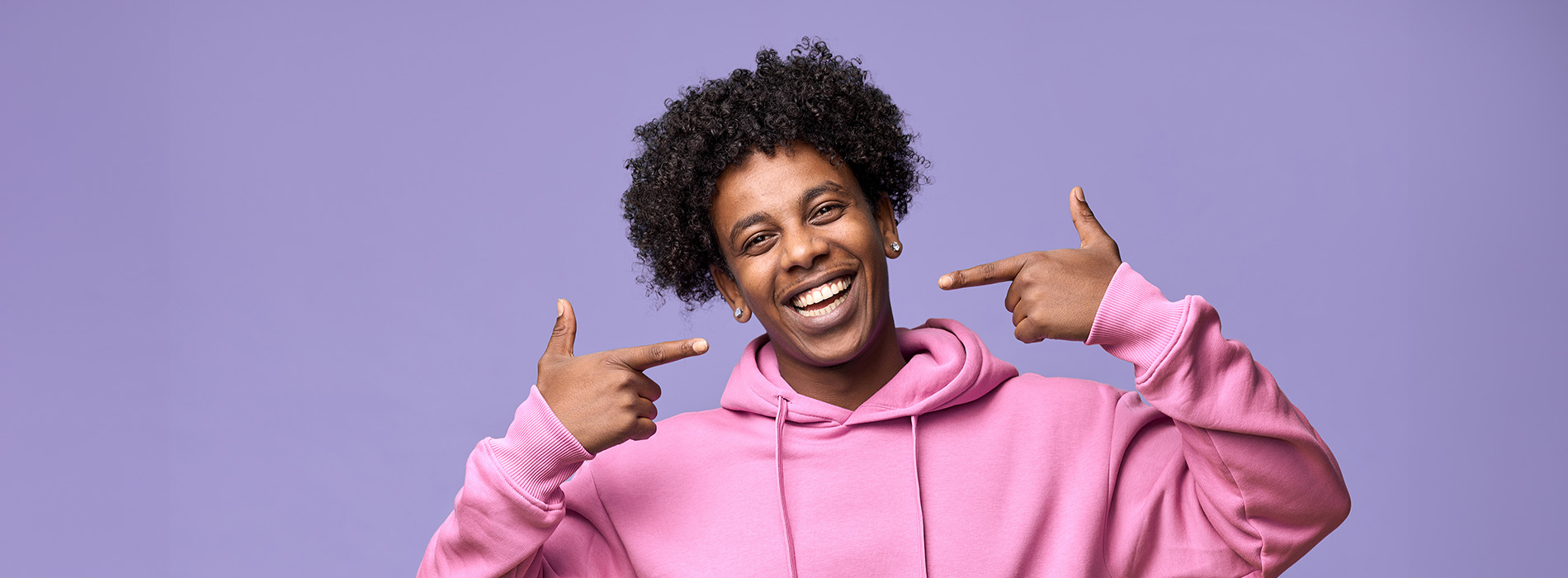 This is a color photograph featuring a person with dark hair wearing a pink hoodie, pointing at their face with both hands, against a purple background.