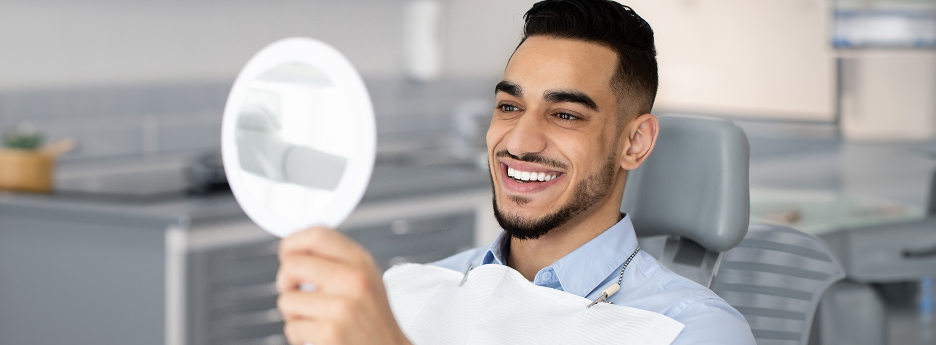 A man sitting at a dental chair, holding up a mirror towards his face with a smile.