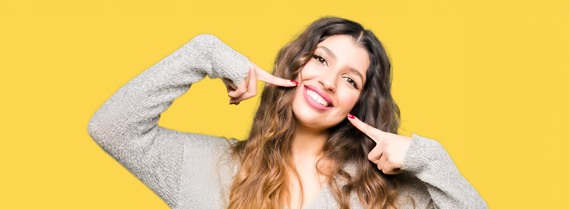 A woman with dark hair is smiling at the camera while making a peace sign gesture with her hand. She is wearing a white sweater and standing against a yellow background.