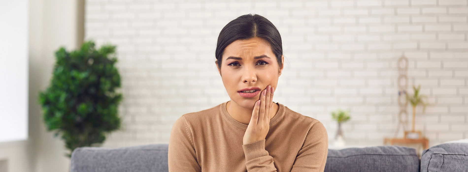 The image shows a woman with her hand on her face, appearing surprised or concerned, standing indoors in front of a couch and a potted plant.