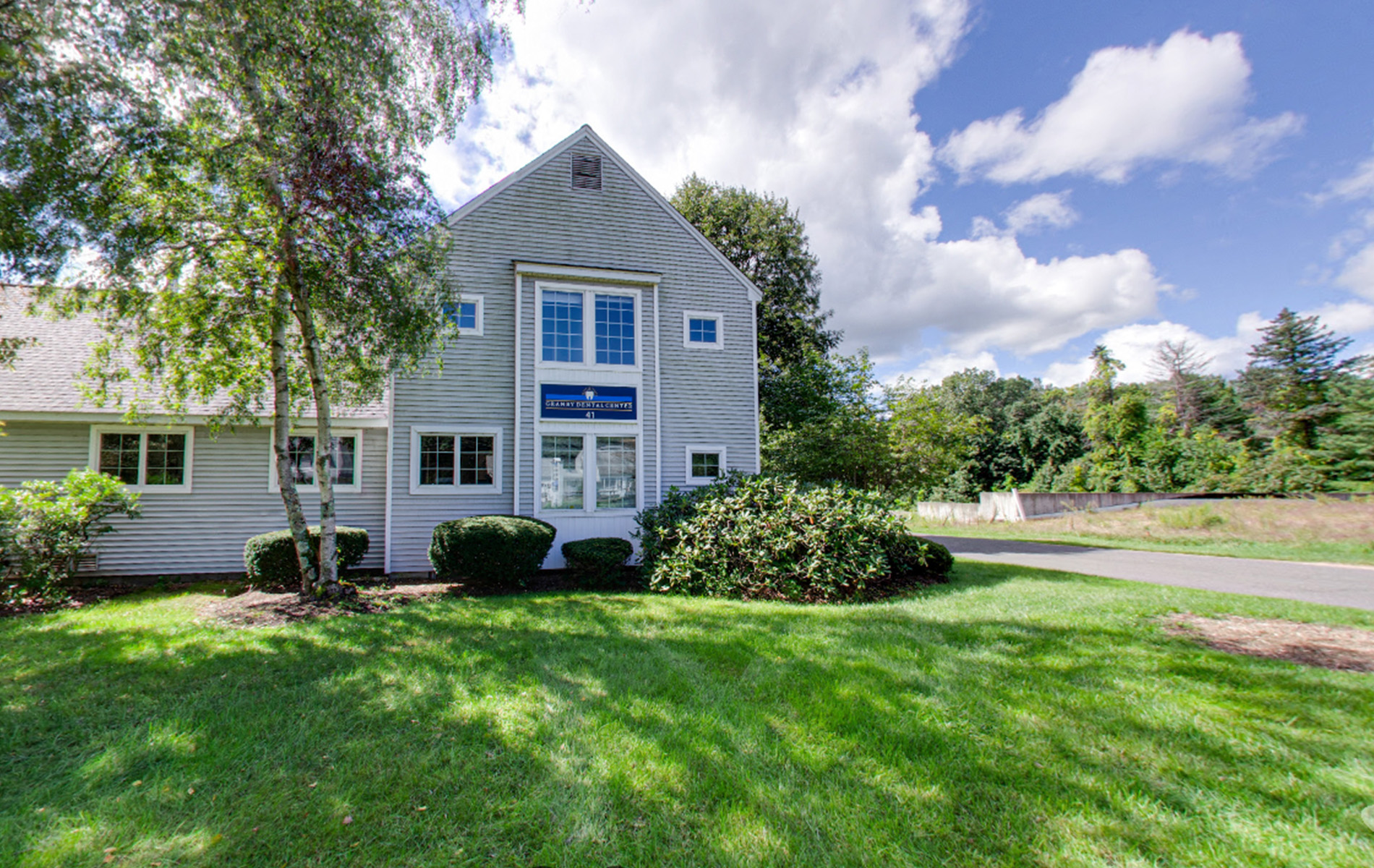 The image shows a two-story house with a white exterior and black shutters, surrounded by a well-maintained lawn under a clear blue sky.