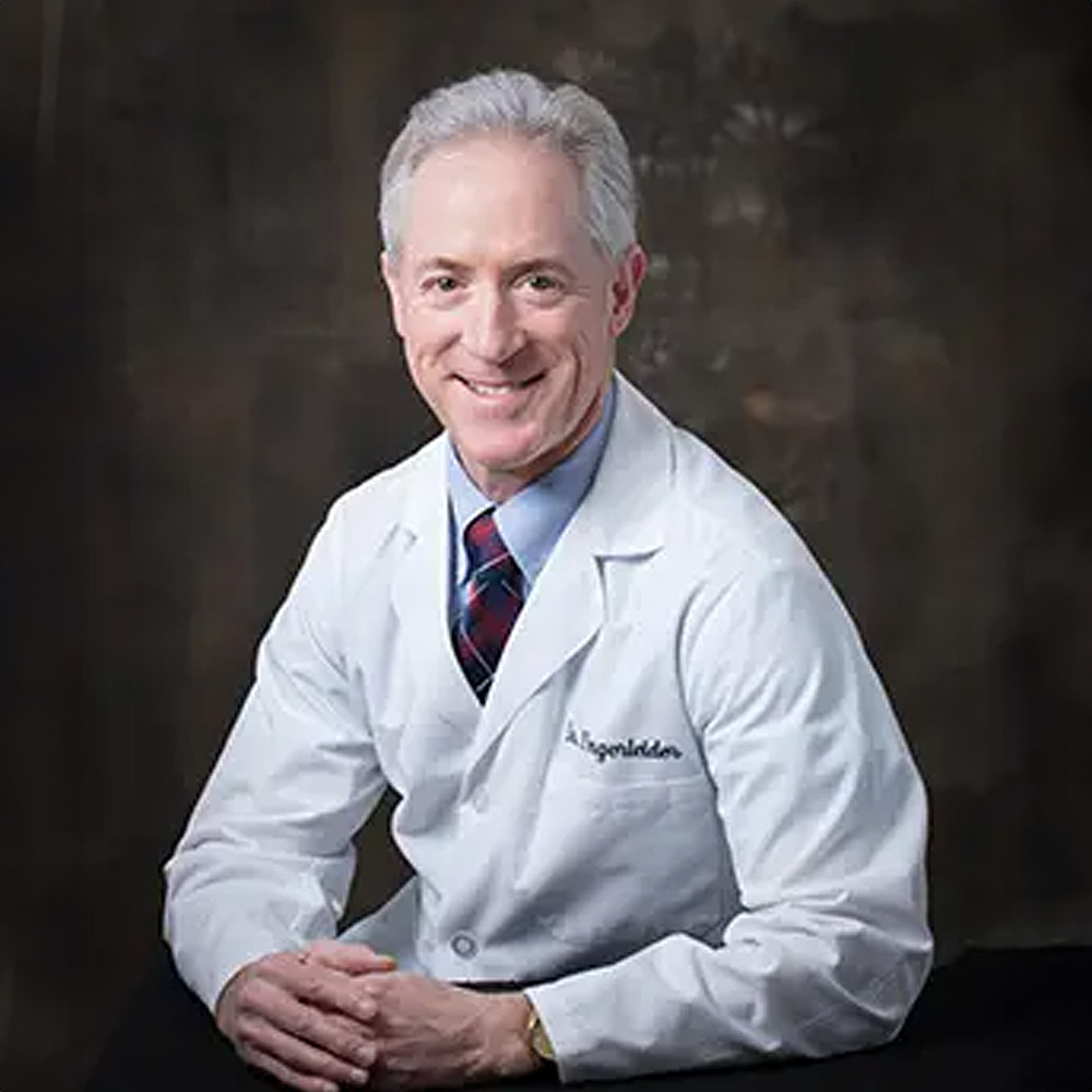 The image shows a man posing for a portrait  he is wearing a white lab coat with name tags and appears to be a professional, possibly a scientist or academic, standing against a dark background with his arms crossed.
