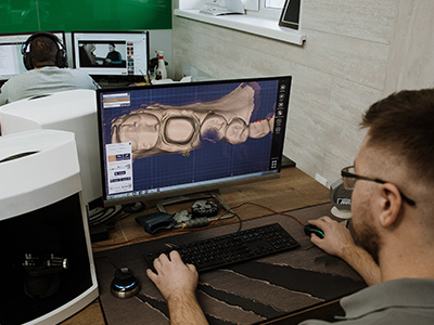 A man sitting at a desk using a computer with a large screen displaying a 3D model of a human skull, surrounded by other computers and equipment indicative of a tech or design workspace.