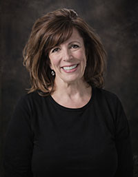 The image shows a woman with short brown hair smiling at the camera, wearing a black top and standing against a blurred background that suggests an indoor setting with lighting equipment.