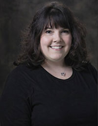 The image shows a woman with short hair smiling at the camera. She has a necklace around her neck and is wearing a black top. Her background is blurred but appears to be an indoor setting with a dark wall.