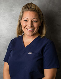 The image features a woman wearing a blue scrub top, standing behind a desk with a nameplate on it, smiling at the camera.