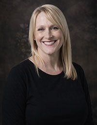 The image shows a woman with blonde hair, wearing a black top, posing for a portrait with a neutral background.
