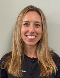 The image shows a woman with long hair smiling at the camera, wearing a black shirt and standing against a white wall.