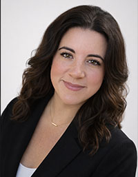 The image features a woman posing for a portrait with a neutral background. She has short hair, wears a black blazer over a white shirt, and has a slight smile on her face.