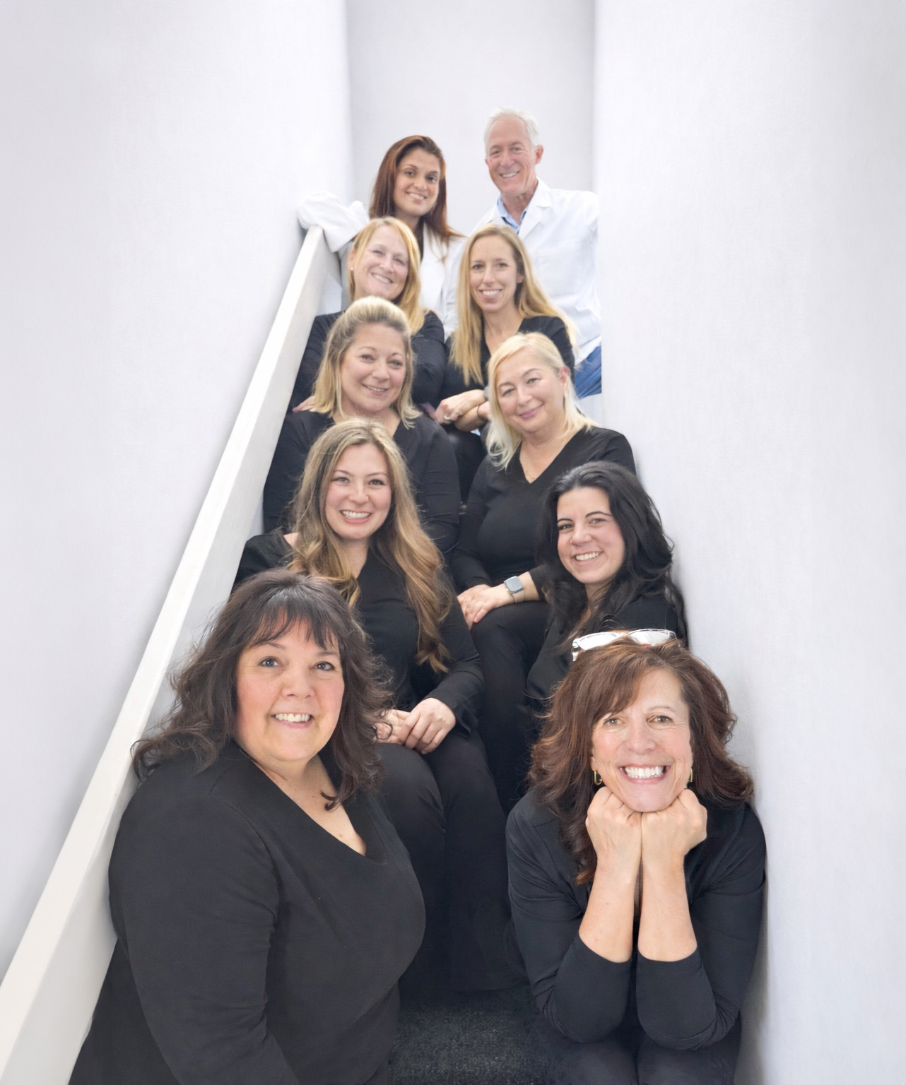 The image shows a group of individuals standing on a staircase, possibly posing for a photograph, with a man standing at the top of the stairs and smiling towards them.