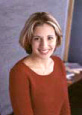A woman with short hair stands against a blurred background, smiling slightly at the camera.