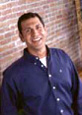 The image shows a man standing indoors, smiling at the camera, wearing a blue shirt with rolled-up sleeves and dark pants. He has short hair and appears to be of middle age. Behind him is a brick wall and a partial view of another person s face in the background.