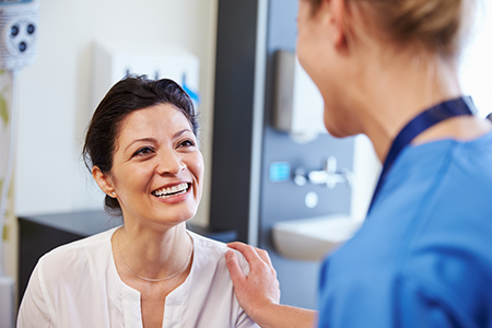 An image featuring two individuals in a medical setting, with one person seated and smiling at another standing individual who appears to be a healthcare professional.