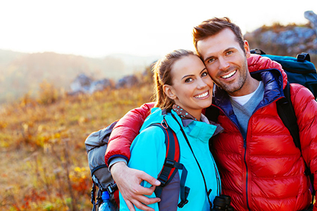 The image shows a man and a woman embracing each other outdoors during daylight, with both dressed for hiking and enjoying a moment together.