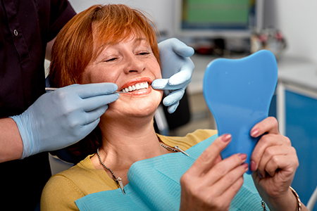 A woman with red hair sitting in a dental chair holding up a blue model tooth, smiling at the camera while being attended to by a dental professional.