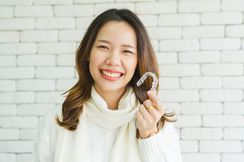 A smiling woman holding a small white object with her right hand, posing against a brick wall background.
