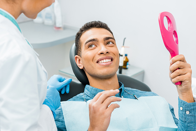 A man is sitting in a dental chair with a smile on his face while holding a pink object, being attended to by a dental professional who is adjusting his mouthpiece.
