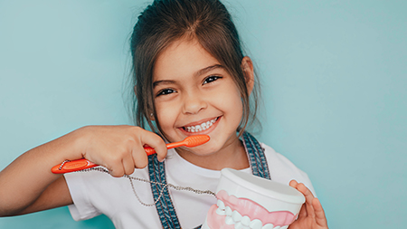 A young girl brushing her teeth with an electric toothbrush while holding a cup of toothpaste.
