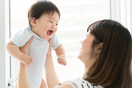 A smiling baby being held by a woman with her arms crossed, both indoors.