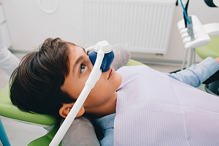 A young person wearing an eye mask sits in a dental chair, looking at the camera with a relaxed expression.