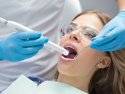 The image shows a person receiving dental care with a toothbrush being held near their mouth by a dental professional wearing blue gloves, who appears to be cleaning or inspecting the teeth.